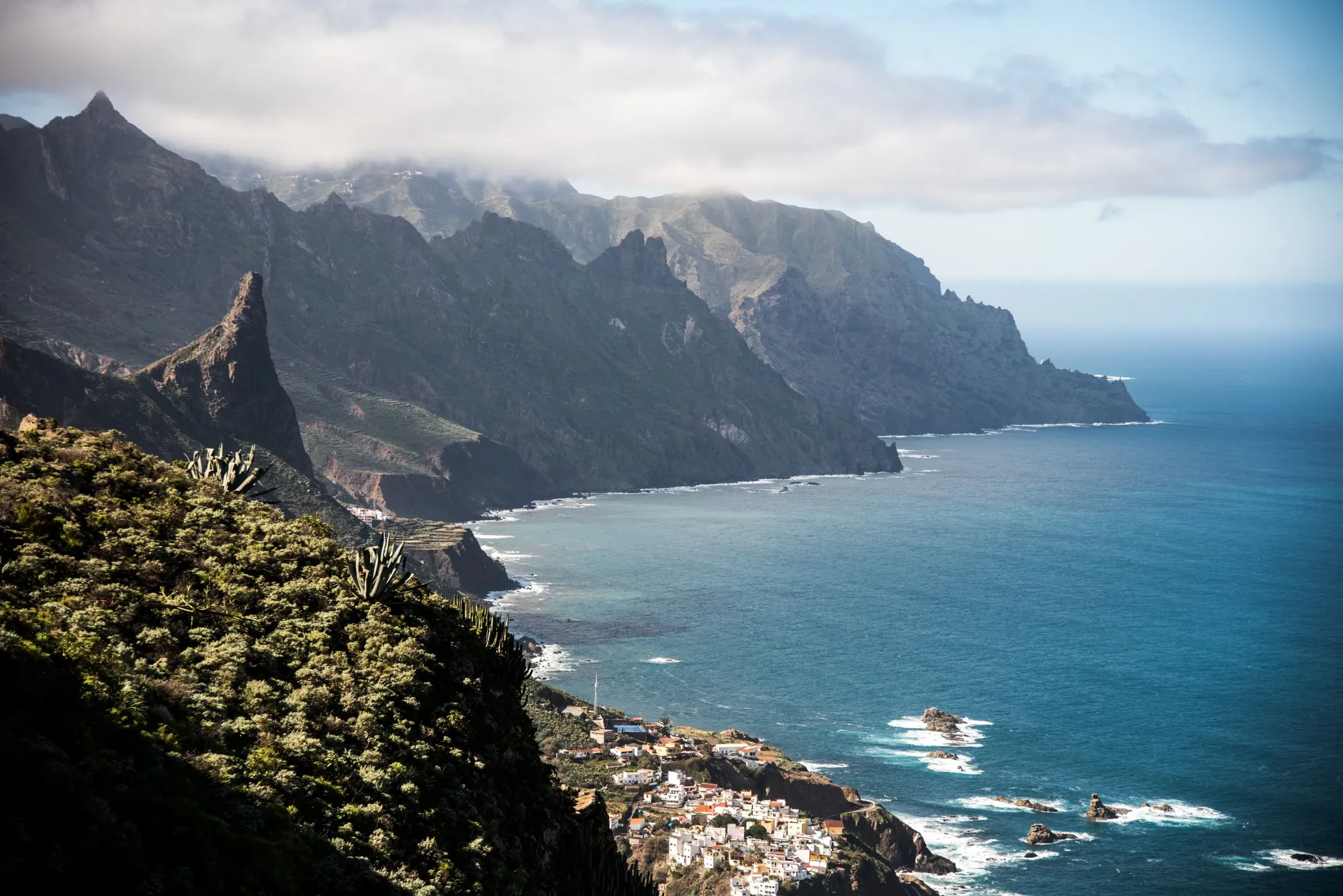 Green mountain ridges of Anaga running down to the Atlantic, with a white village on a hillside and a coast road below