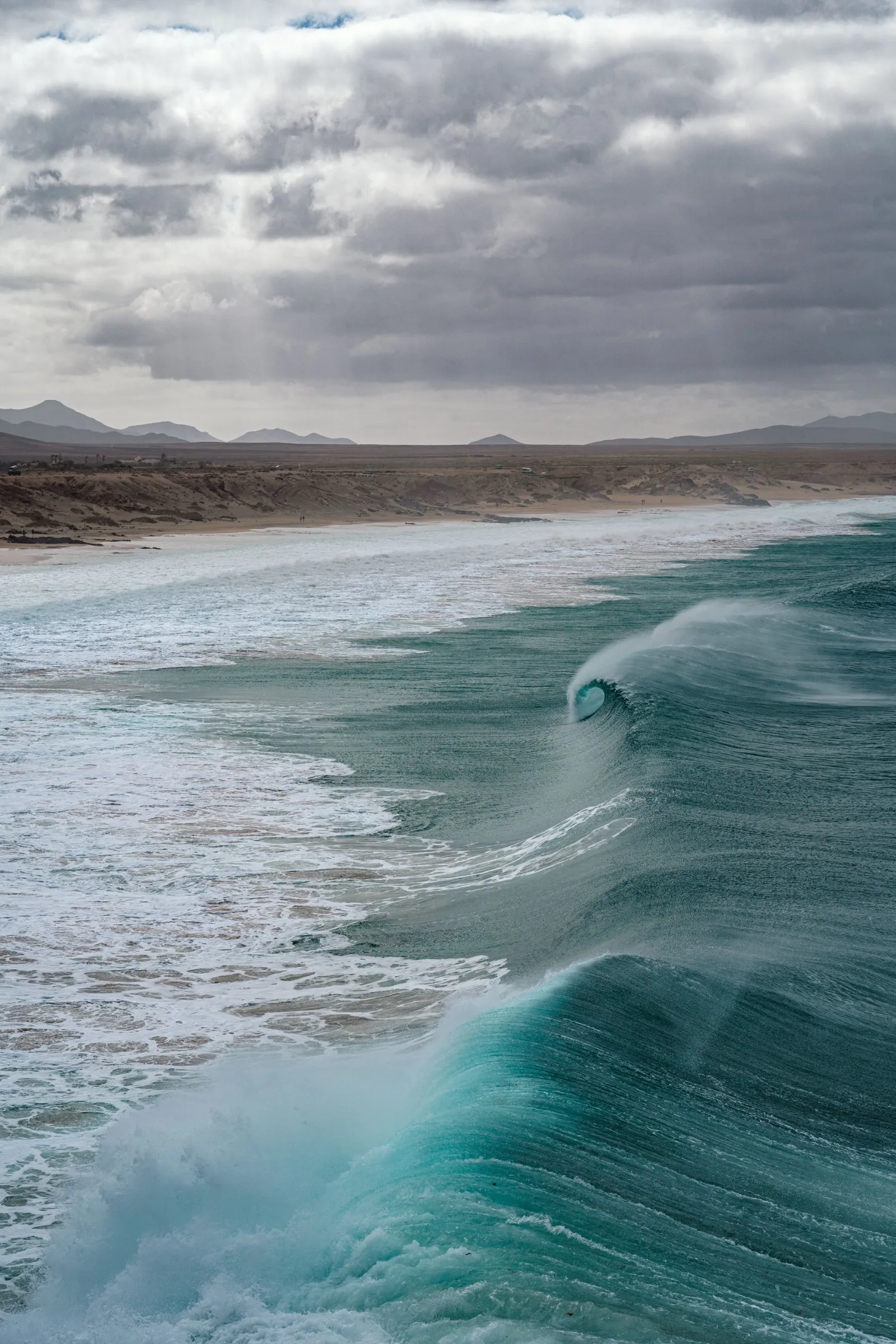 A turquoise wave curling towards a sandy beach under grey clouds