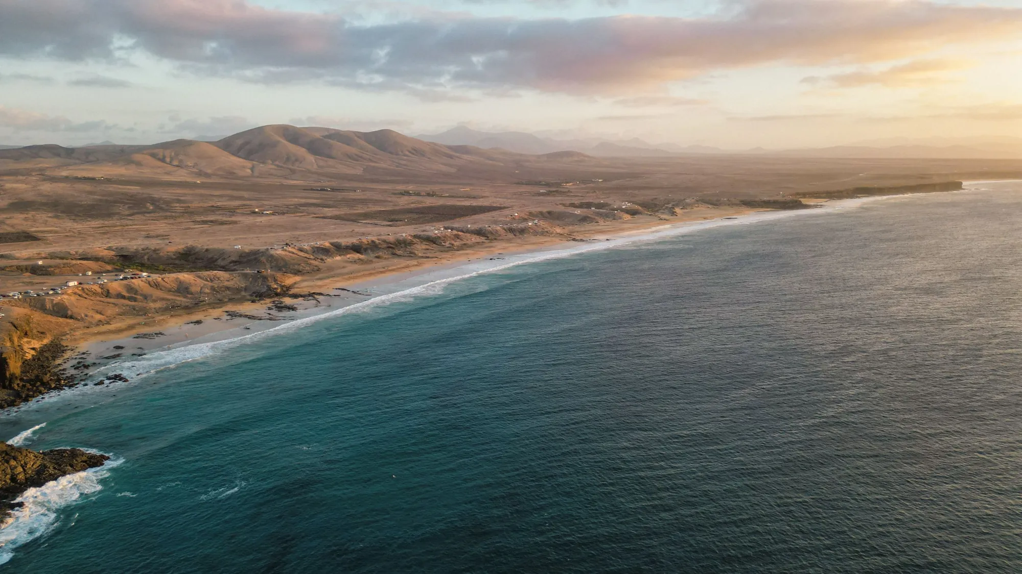 A long beach and turquoise sea below brown hills at sunset on Fuerteventura